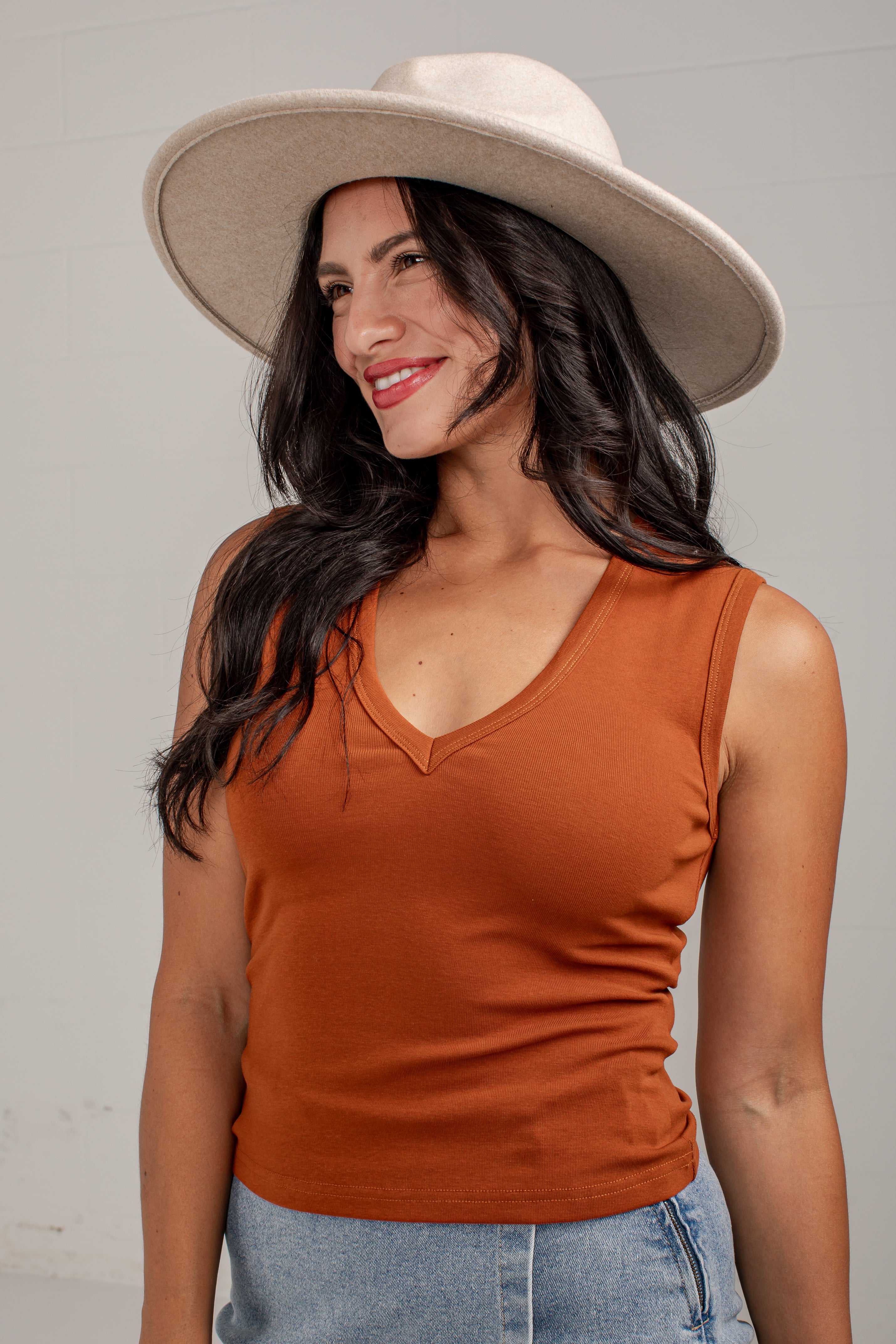 A woman wearing a sleeveless burnt orange V-neck tank top, a wide-brimmed cream hat, and light blue jeans, photographed against a light gray studio background.