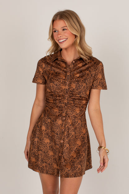 A woman modeling a short-sleeve brown and black leopard-print mini dress with a button-up front and gathered waist, photographed against a plain light gray studio background.