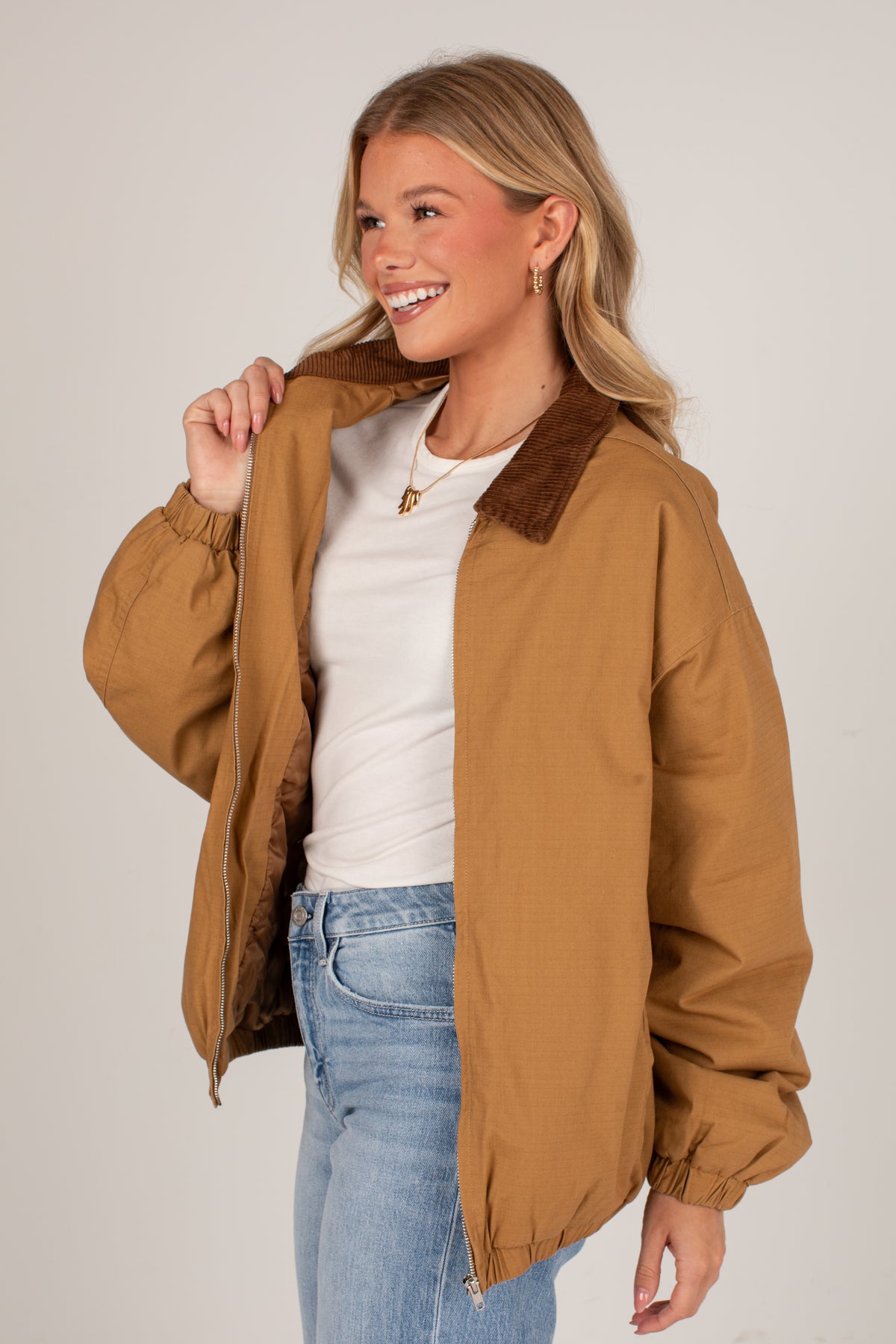 A woman wearing a tan utility-style jacket with a brown corduroy collar, layered over a white t-shirt and blue jeans, posing against a light gray studio background.