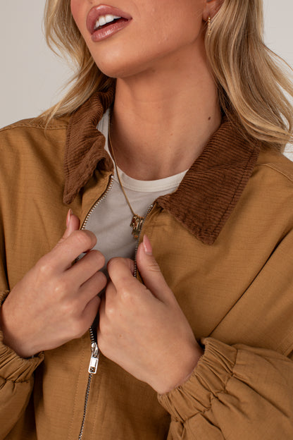 A close-up of a woman wearing a tan zip-up jacket with a brown corduroy collar, zipped over a white t-shirt.