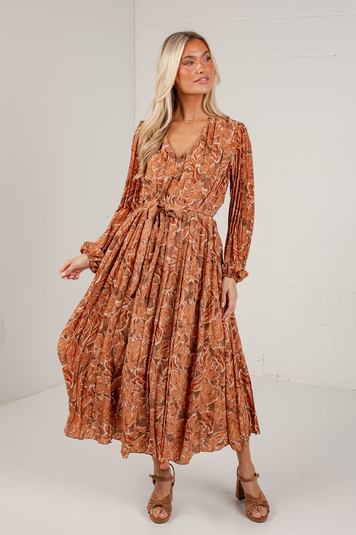 A woman modeling a rust-orange long-sleeve midi dress with a brown abstract floral pattern, standing against a white brick wall.