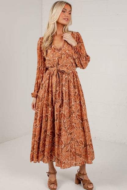 A woman modeling a rust-orange long-sleeve midi dress with a brown abstract floral pattern, featuring a tie waist and puffed sleeves, set against a white brick wall background.