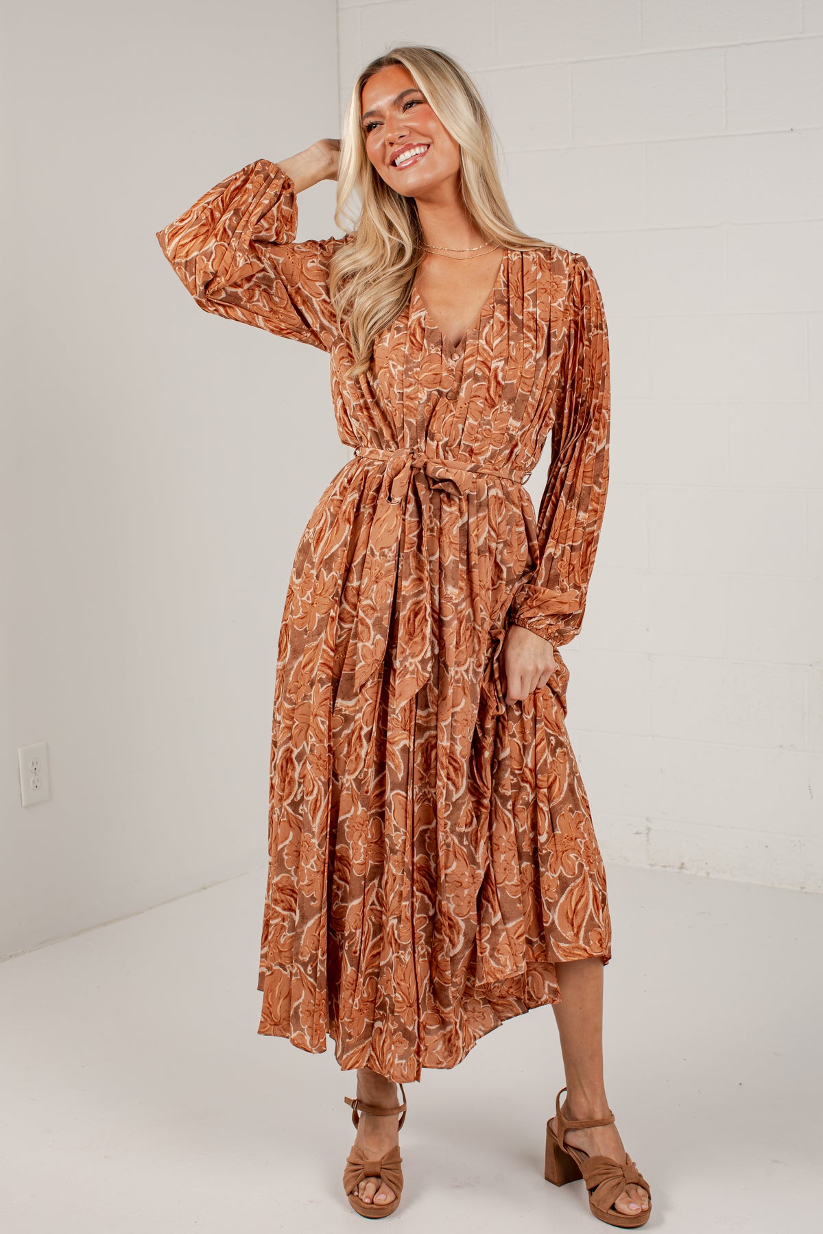 A woman modeling a rust-orange long-sleeve midi dress with a white abstract floral pattern, featuring a tie waist and V-neckline, set against a white studio background.