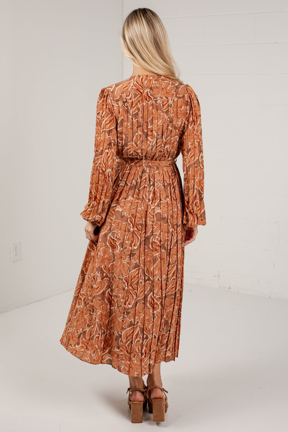 A woman viewed from behind wearing a long-sleeve midi dress with a brown and beige botanical leaf pattern, paired with brown platform sandals, set against a white studio background.