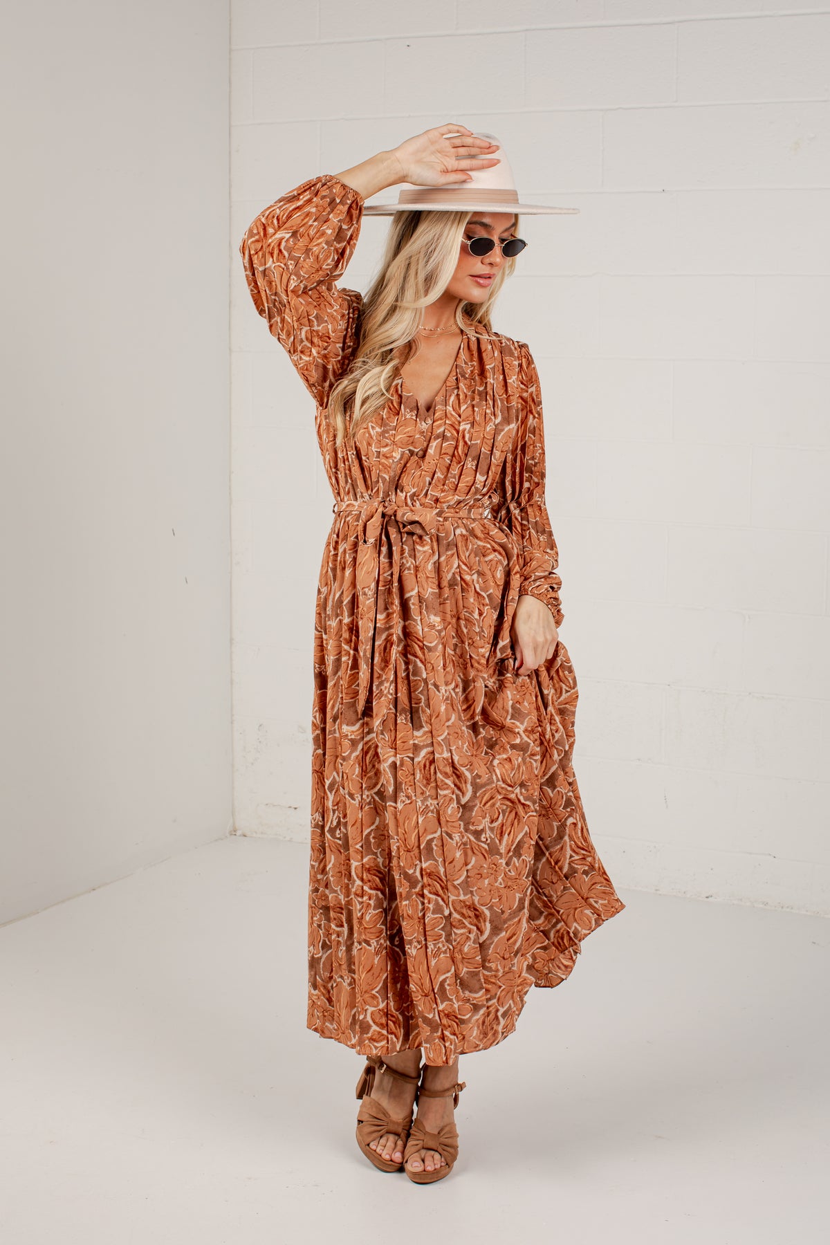 A woman modeling a rust-orange long-sleeve maxi dress with a brown abstract floral pattern, styled with a wide-brimmed straw hat, sunglasses, and brown strappy heels against a white studio background.