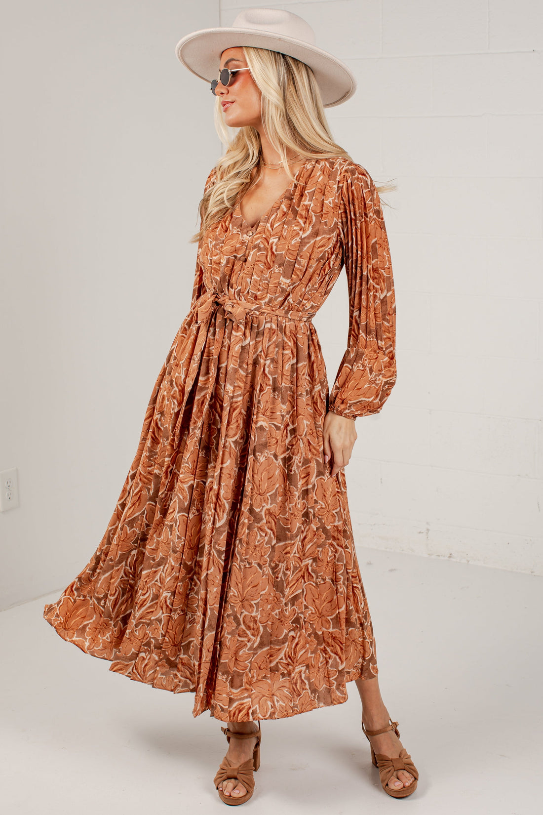 A woman modeling a long-sleeve rust-orange maxi dress with a brown abstract leaf pattern, styled with a beige wide-brim hat, sunglasses, and tan strappy sandals against a white studio background.