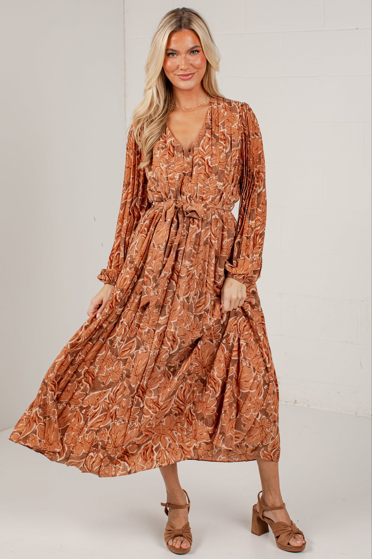 A woman modeling a long-sleeve rust-orange midi dress with a white abstract leaf pattern, featuring a V-neckline and a self-tie waist belt. She is wearing brown block-heel sandals against a white studio background.
