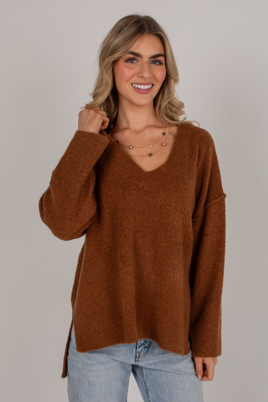A smiling blonde woman wearing a fuzzy brown V-neck sweater and light blue jeans, standing against a plain light gray studio background.