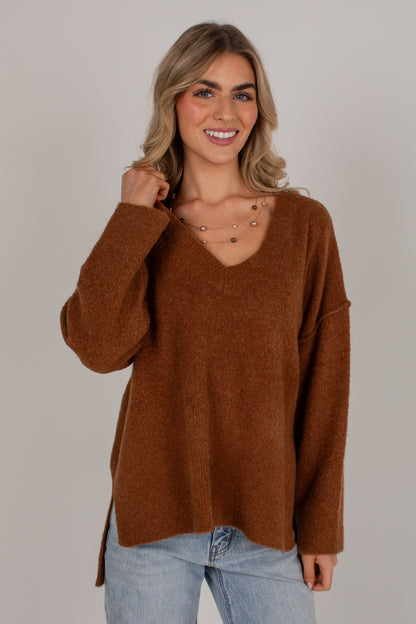 A smiling blonde woman wearing a fuzzy brown V-neck sweater and light blue jeans, standing against a plain light gray studio background.
