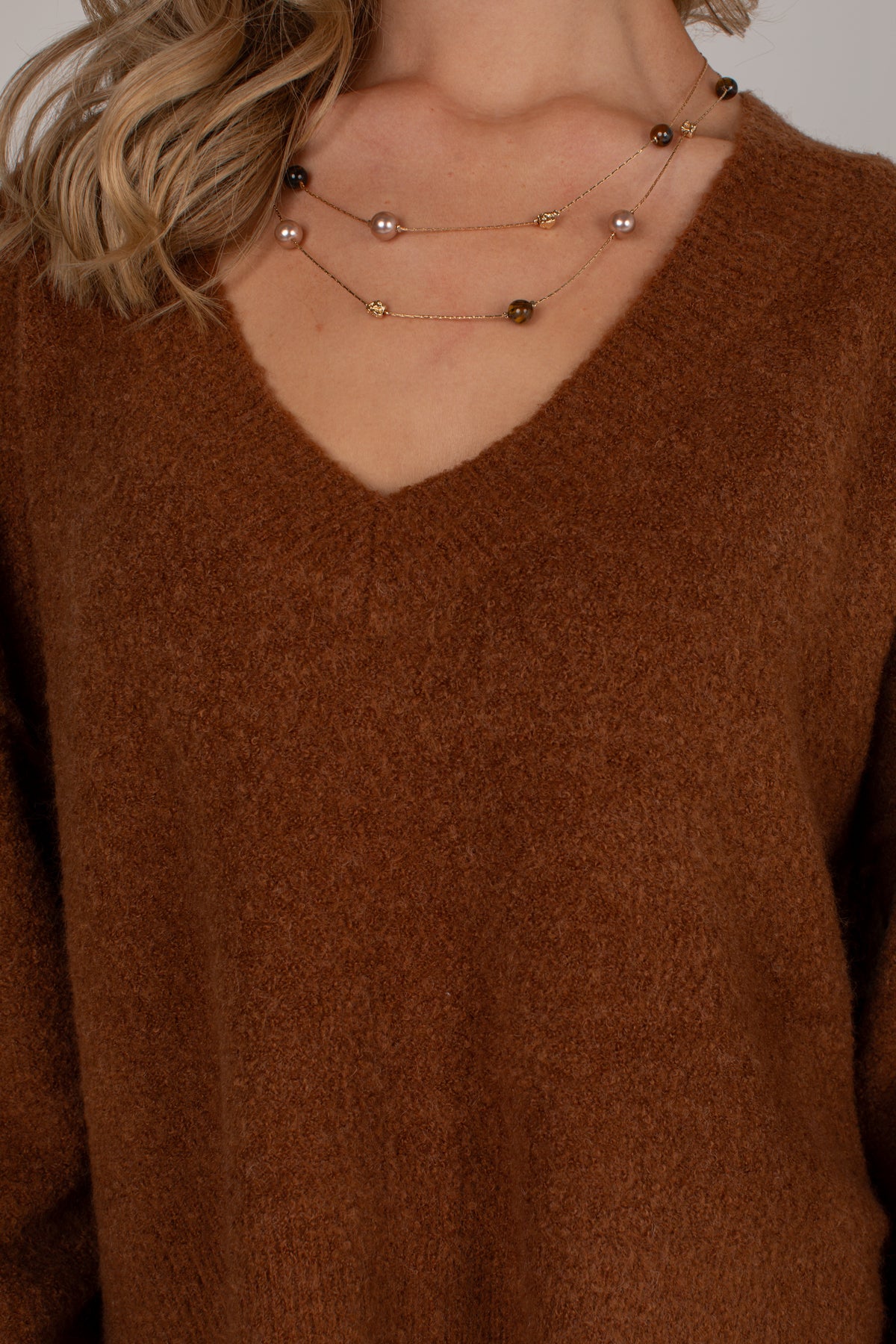 A close-up of a person wearing a brown V-neck knit sweater and layered gold necklaces with pearl-like beads.