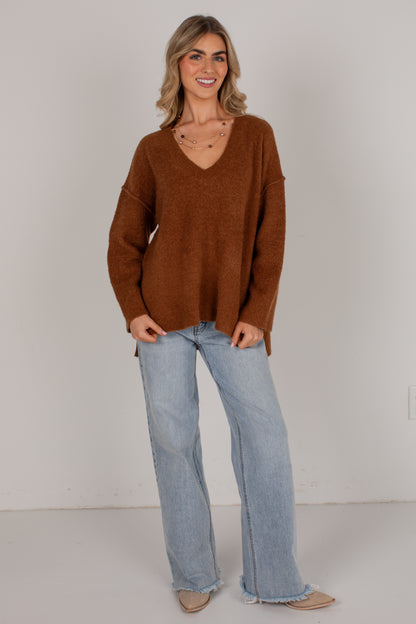 A woman modeling an oversized brown knit sweater with a V-neck, paired with light-wash wide-leg jeans and beige flats, standing against a plain off-white studio wall.