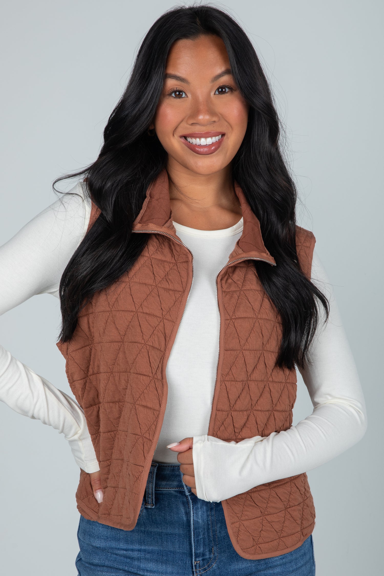 Woman wearing a brown quilted vest over a white long-sleeve shirt and blue jeans on a gray background