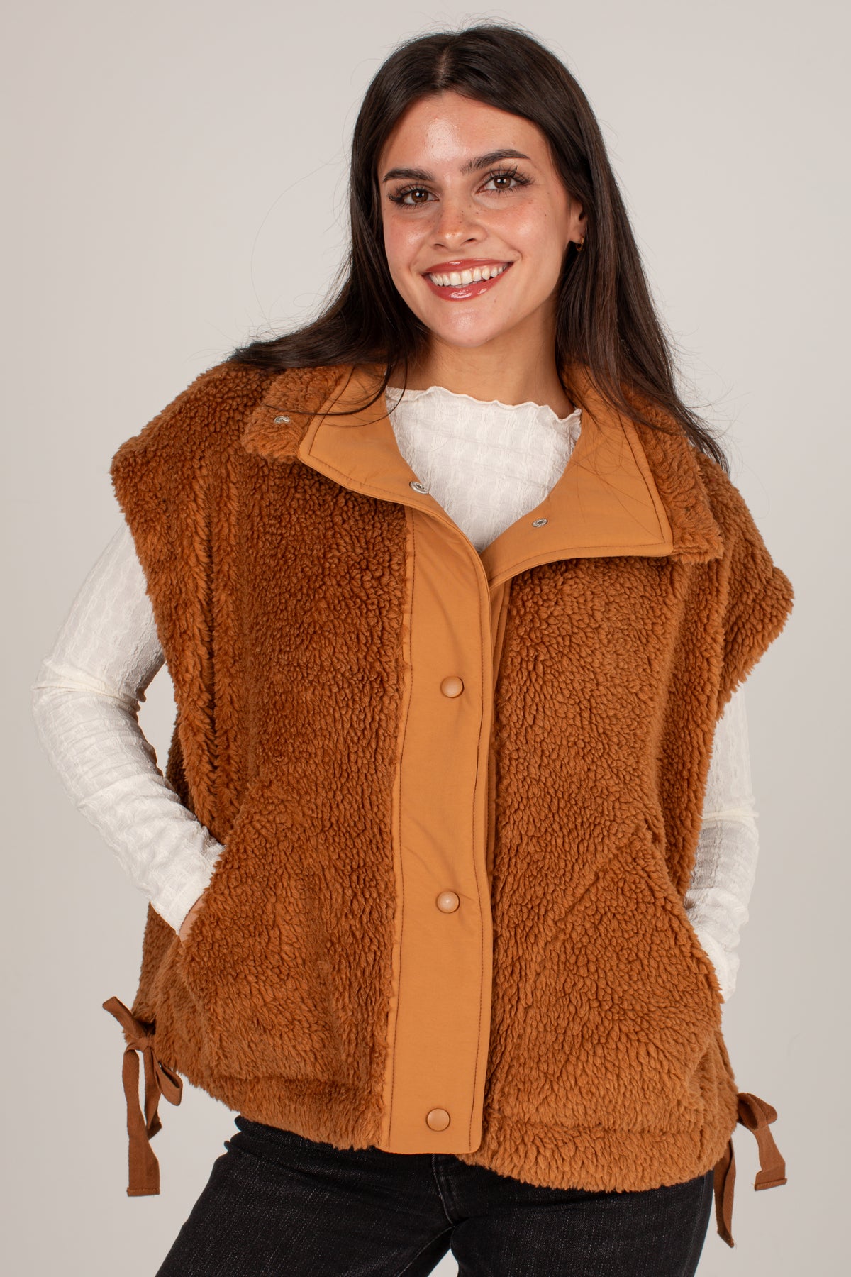 A smiling woman wearing a brown sherpa fleece vest over a white long-sleeve top and black pants, photographed against a plain light gray studio background.
