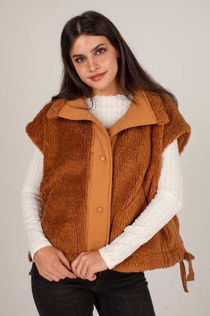 A woman wearing a brown sherpa fleece vest over a white ribbed long-sleeve top and black jeans, photographed against a plain light gray studio background.