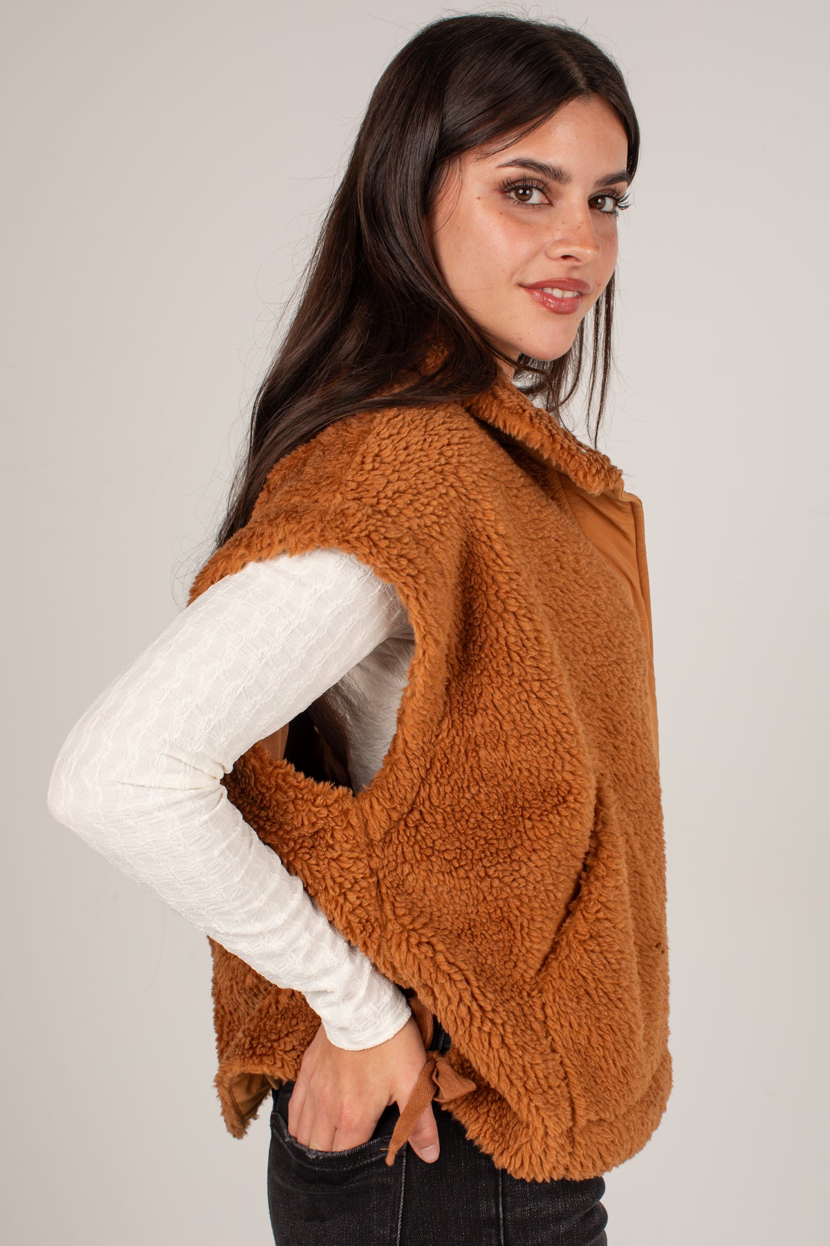 A woman wearing a brown sherpa fleece vest over a white long-sleeve top and black jeans, photographed against a plain light gray studio background.