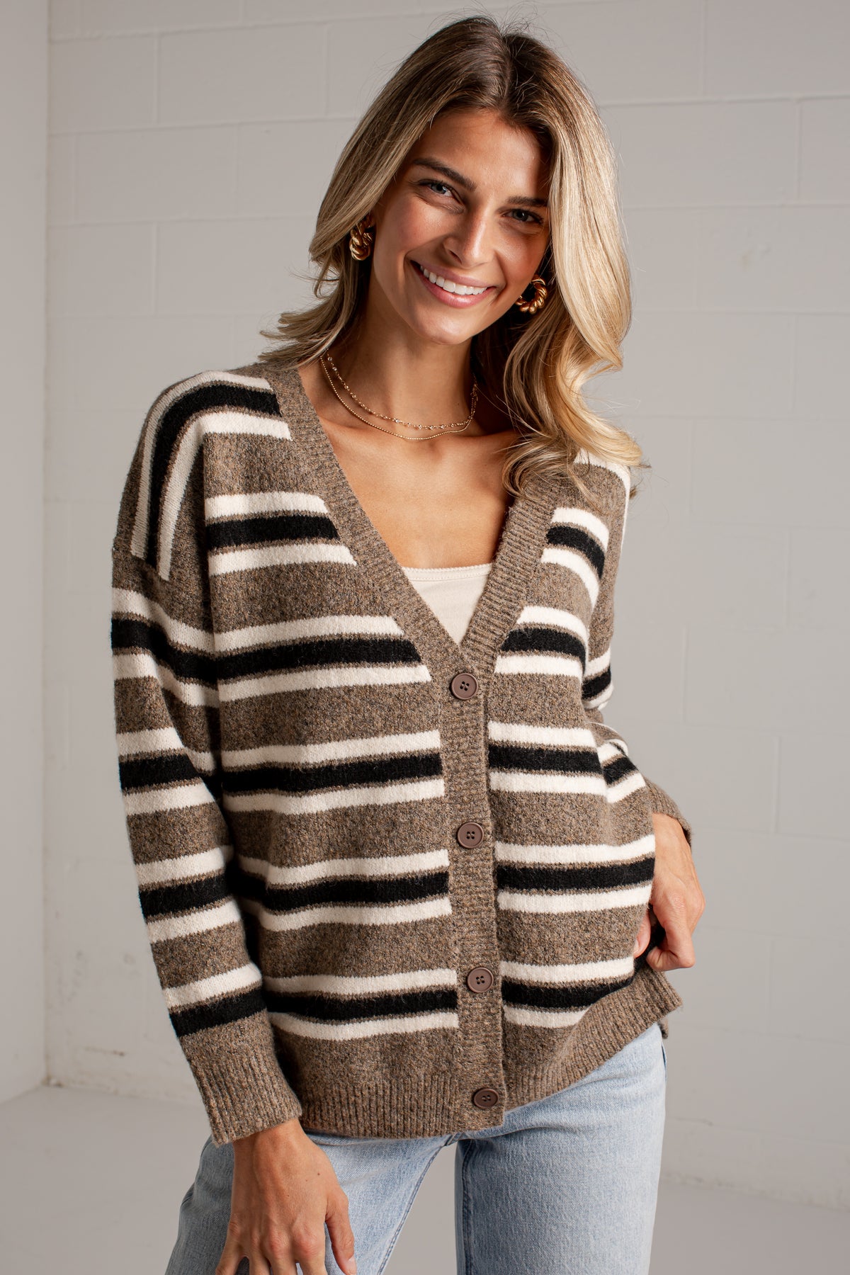 A woman wearing a brown, beige, and black striped knit cardigan sweater with brown buttons, paired with light blue jeans, standing against a white brick wall.