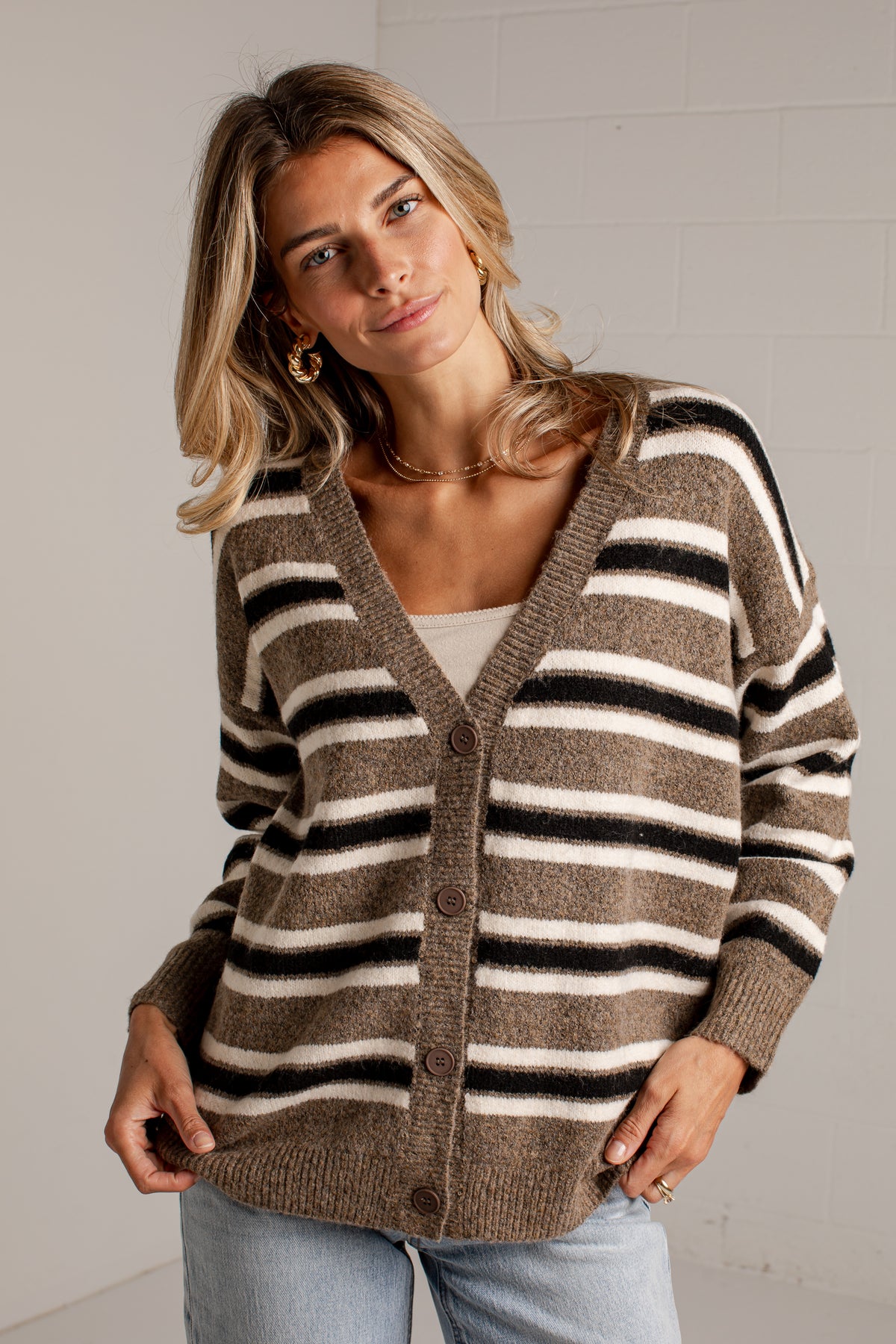 A woman wearing a brown, beige, and black striped knit cardigan sweater over a white top and light blue jeans, standing against a white brick wall.