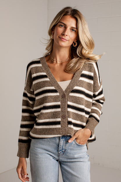A woman wearing a brown, beige, and black striped knit cardigan over a white top and light blue jeans, standing against a light gray brick wall.