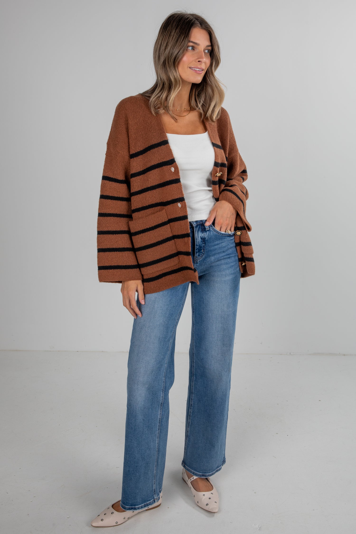 Woman wearing a brown striped cardigan, white top, and blue jeans on a gray background