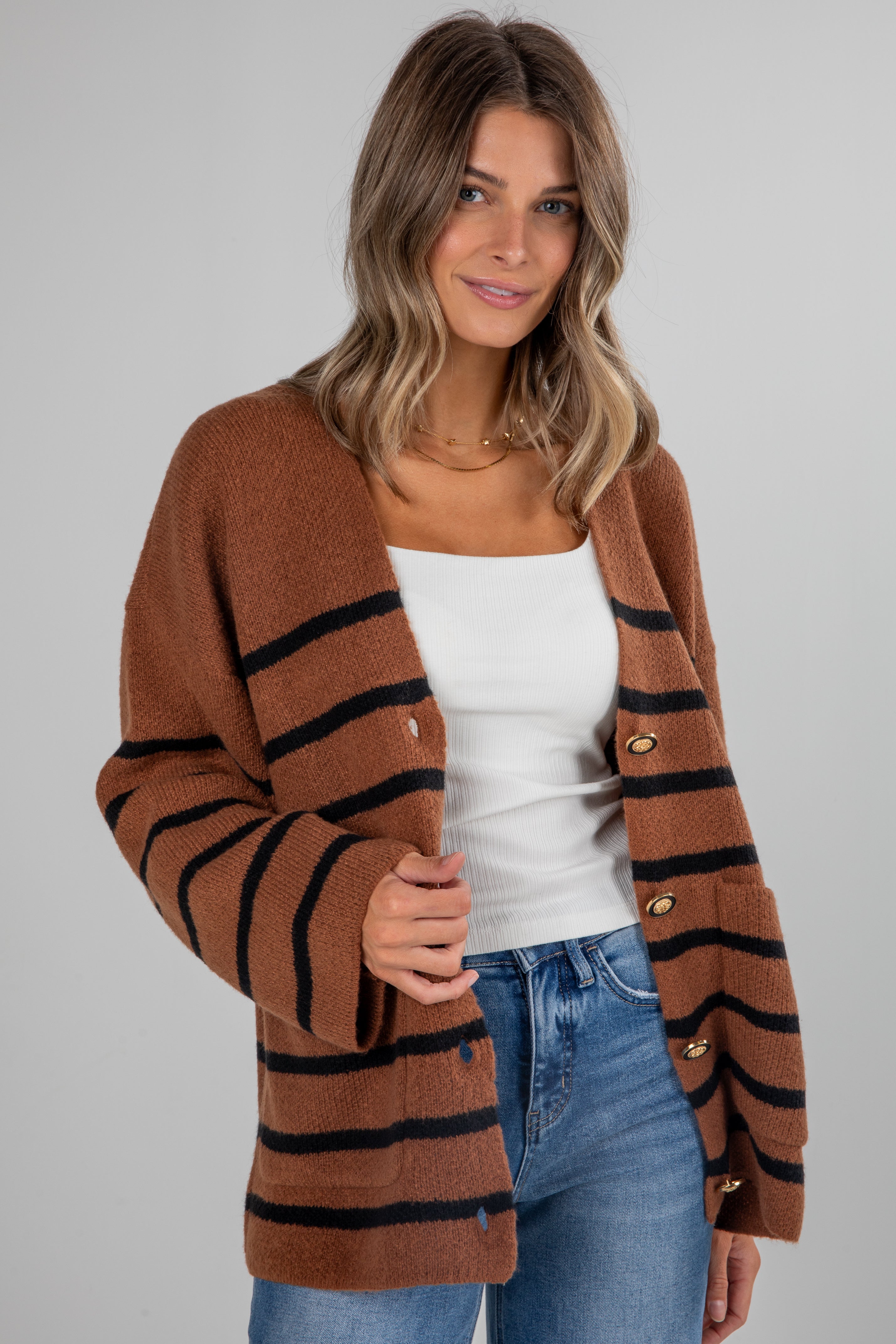 Woman wearing a brown cardigan with black stripes, white top, and blue jeans on a plain background