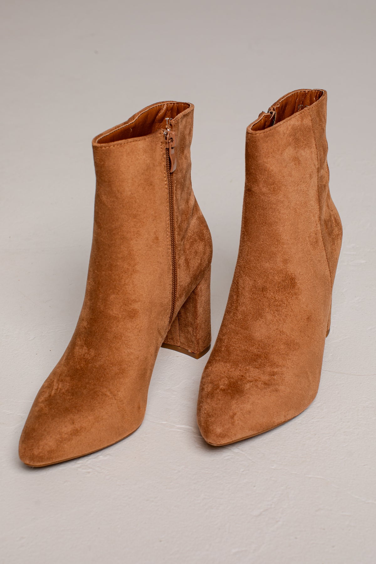A pair of tan suede ankle boots with block heels and side zippers, displayed on a light gray studio background.