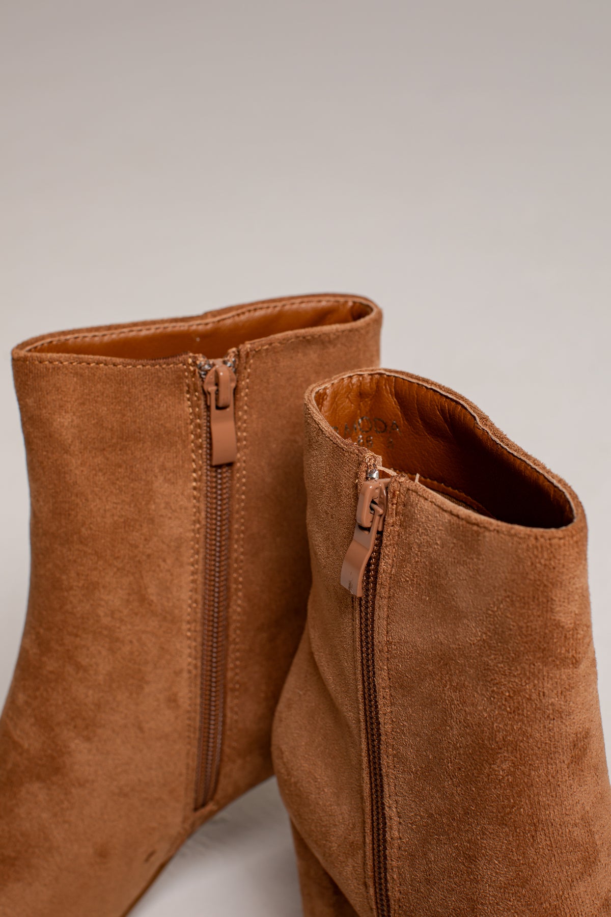 A close-up of a pair of tan suede ankle boots with side zippers, shown against a neutral light gray background.