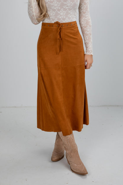 Person wearing a brown suede skirt with a white top and beige boots on a light gray background