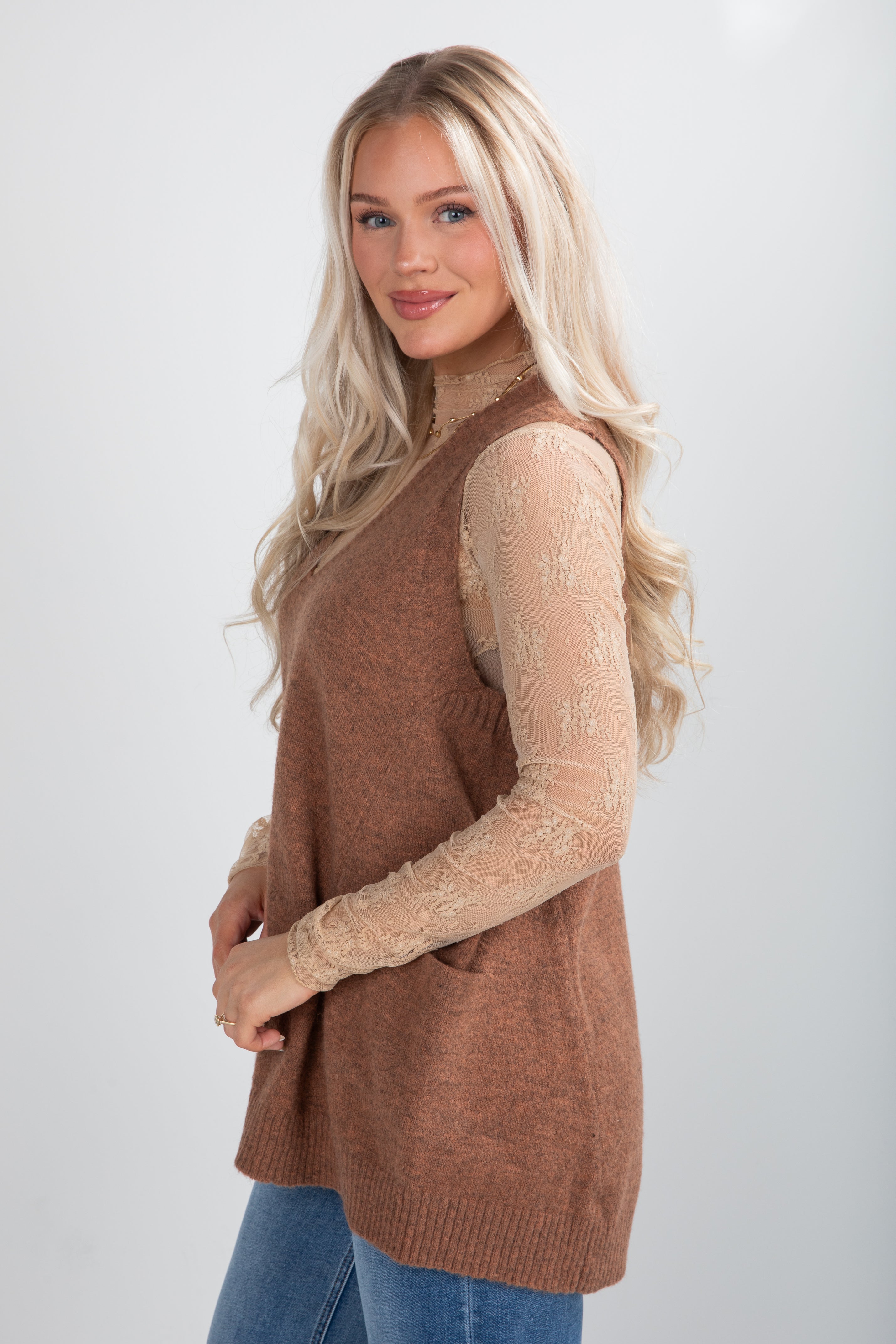 Woman wearing a brown vest over a sheer top with blue jeans on a white background