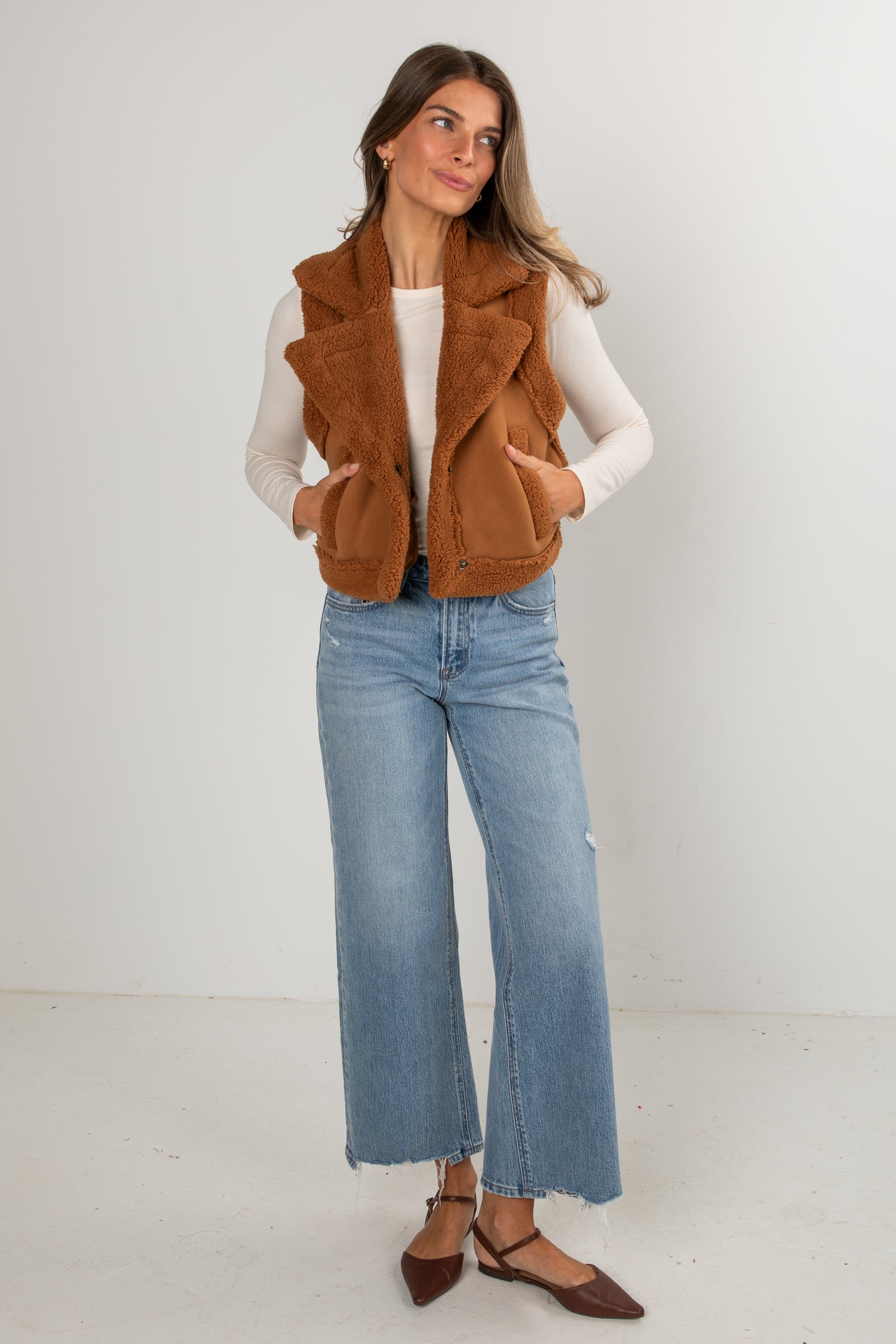 Woman wearing a brown vest, white shirt, and blue jeans against a white background