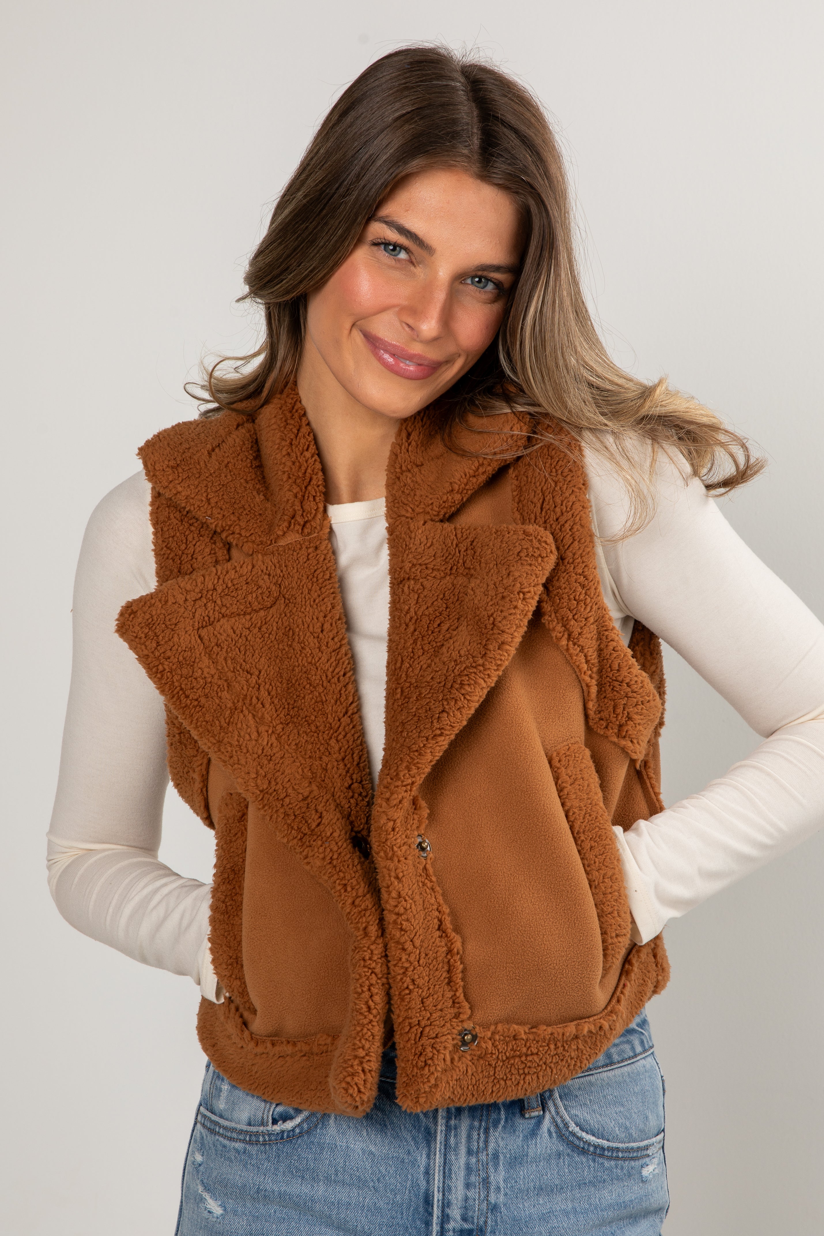 Woman wearing a brown fleece vest over a white long-sleeve shirt and blue jeans on a plain background