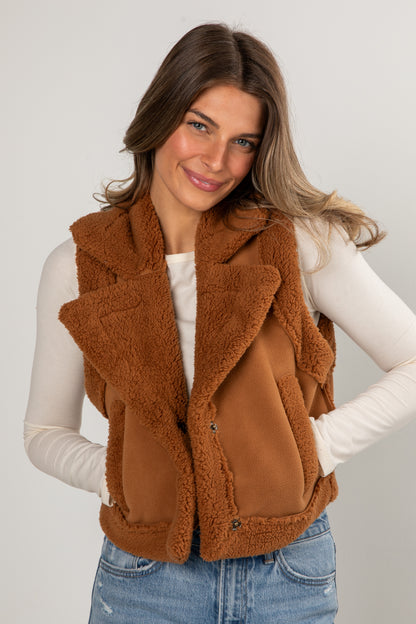 Woman wearing a brown fleece vest over a white long-sleeve shirt and blue jeans on a plain background