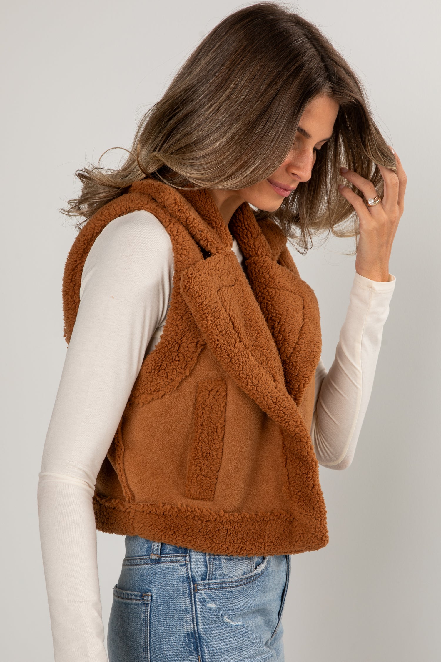 Woman wearing a brown fleece vest over a white long-sleeve shirt and blue jeans on a plain background