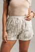 A close-up of a person wearing light beige and gray patterned drawstring shorts with a beige top, shown against a plain light gray background.