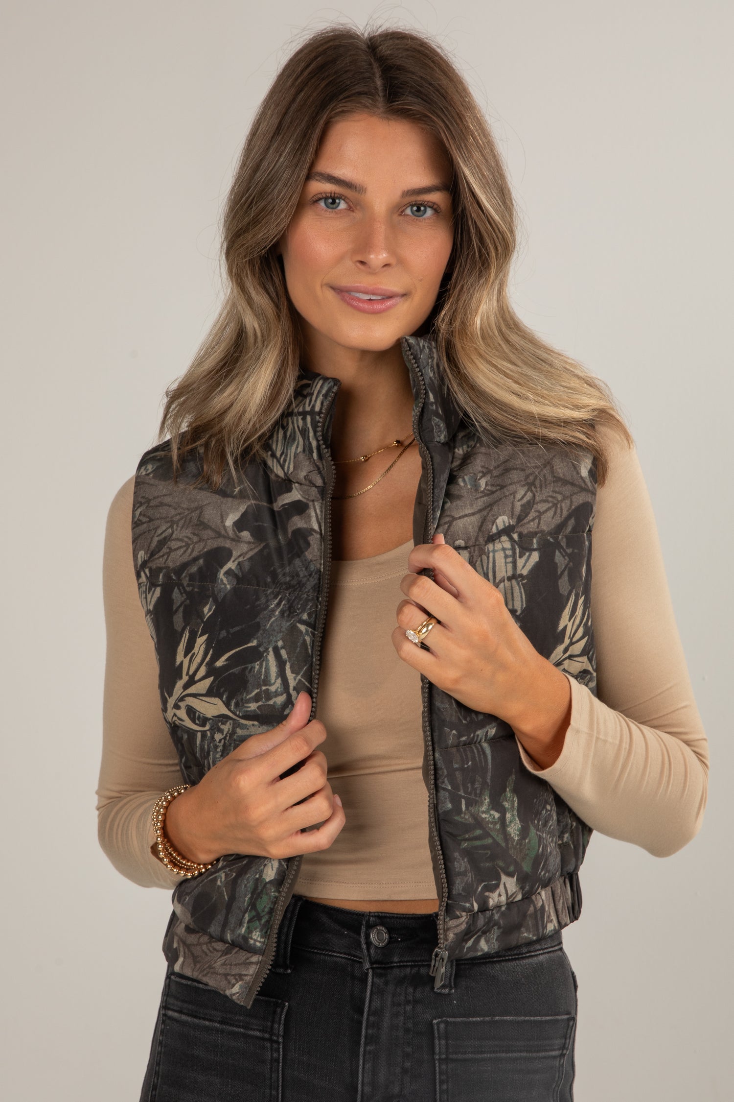 Woman wearing a camouflage vest over a beige shirt on a plain background