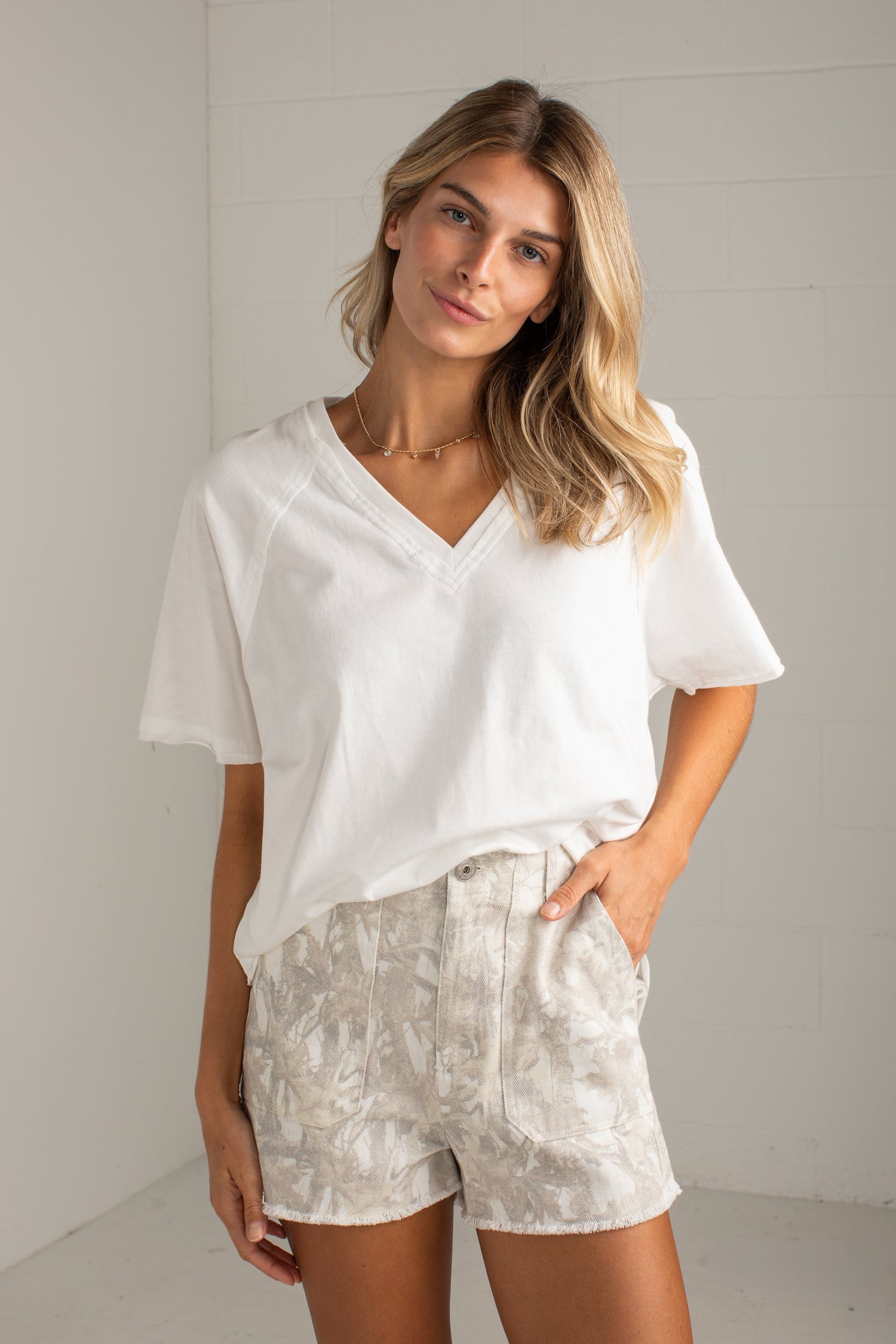 A woman modeling a white short-sleeve V-neck top and light beige patterned shorts against a white brick wall background.