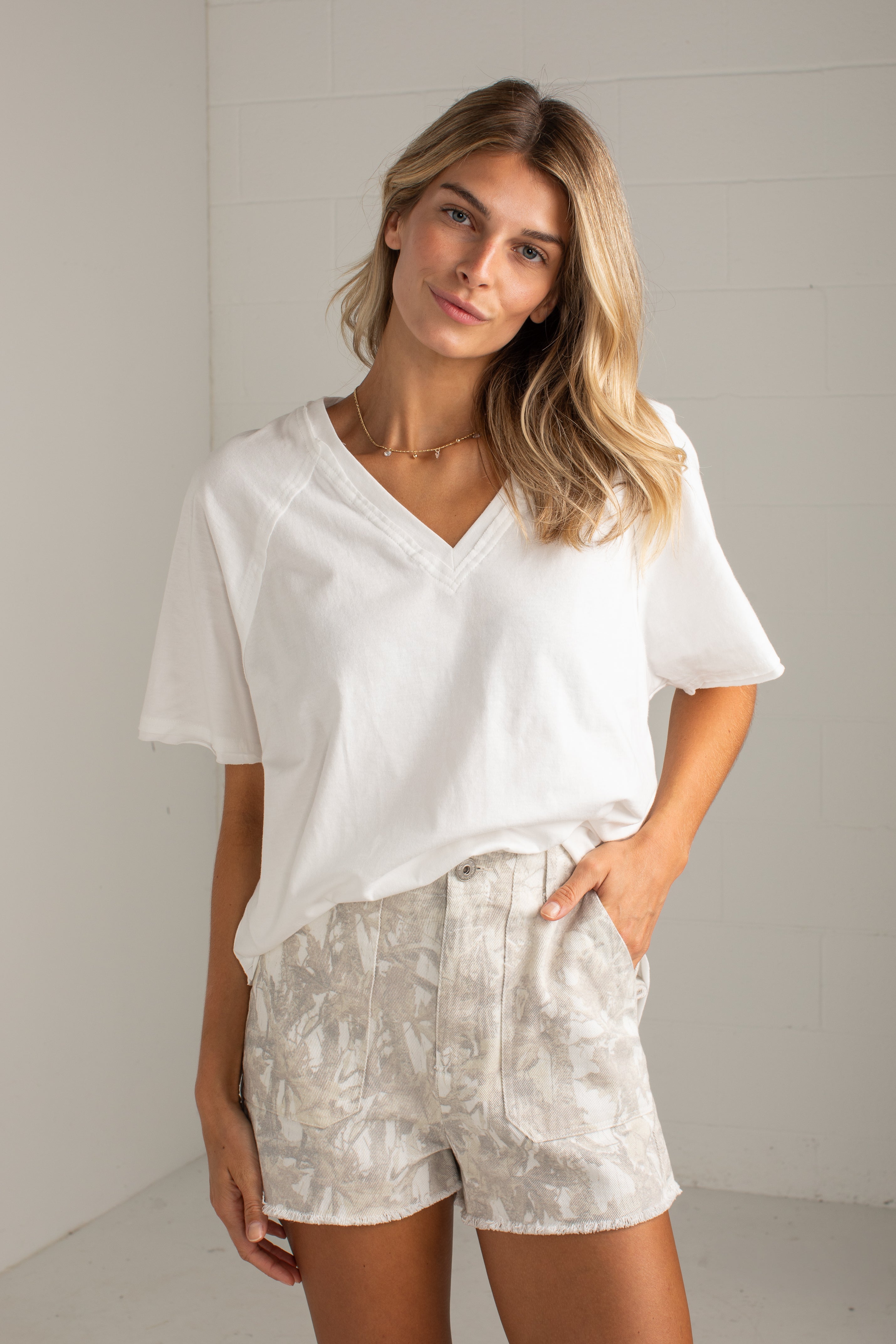 A woman modeling a white short-sleeve V-neck top and light beige patterned shorts against a white brick wall background.
