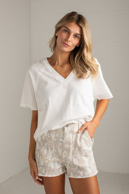 A woman modeling a white short-sleeve V-neck top and light beige patterned shorts against a white brick wall background.