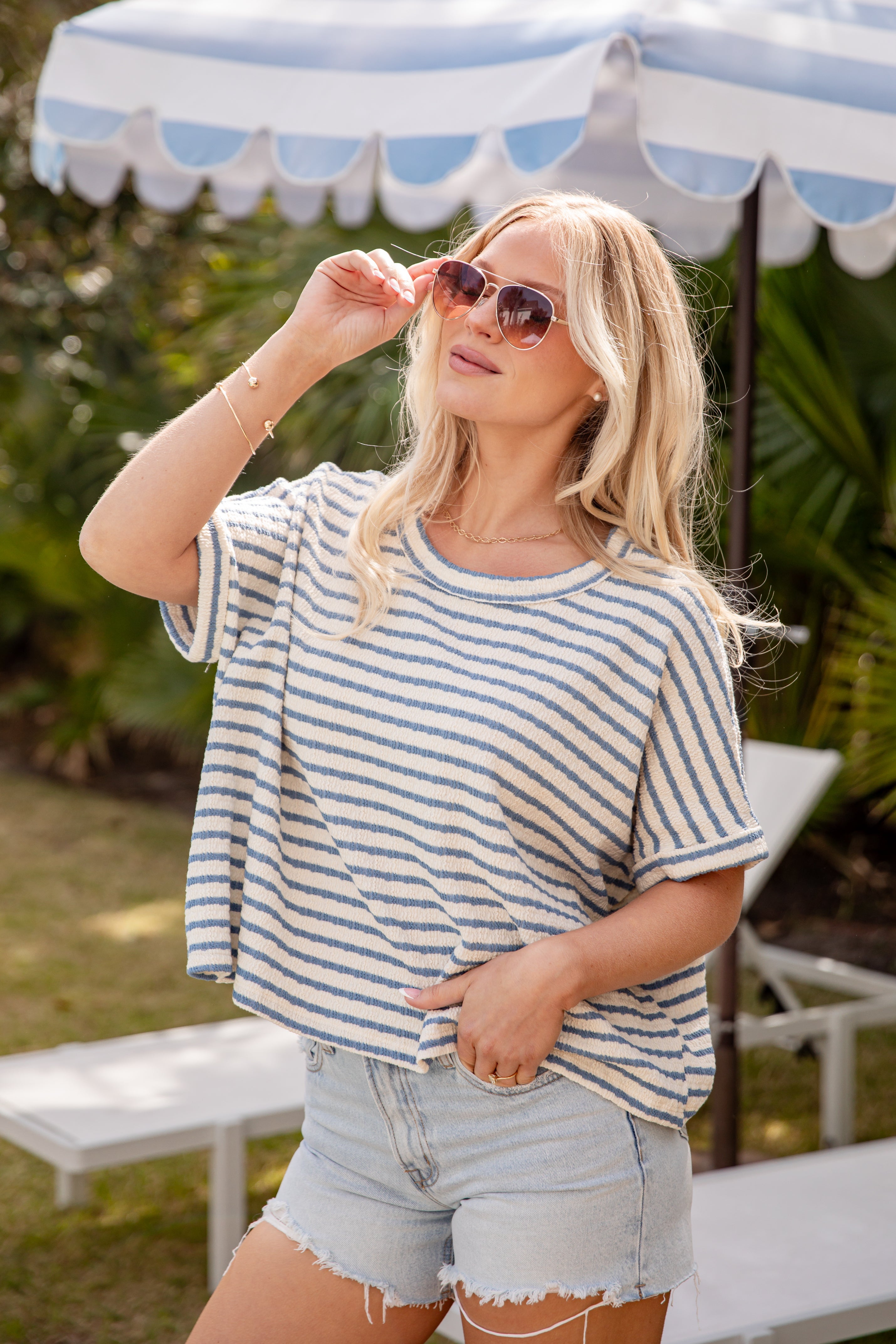 Woman wearing a striped shirt and shorts under a striped umbrella with greenery in the background