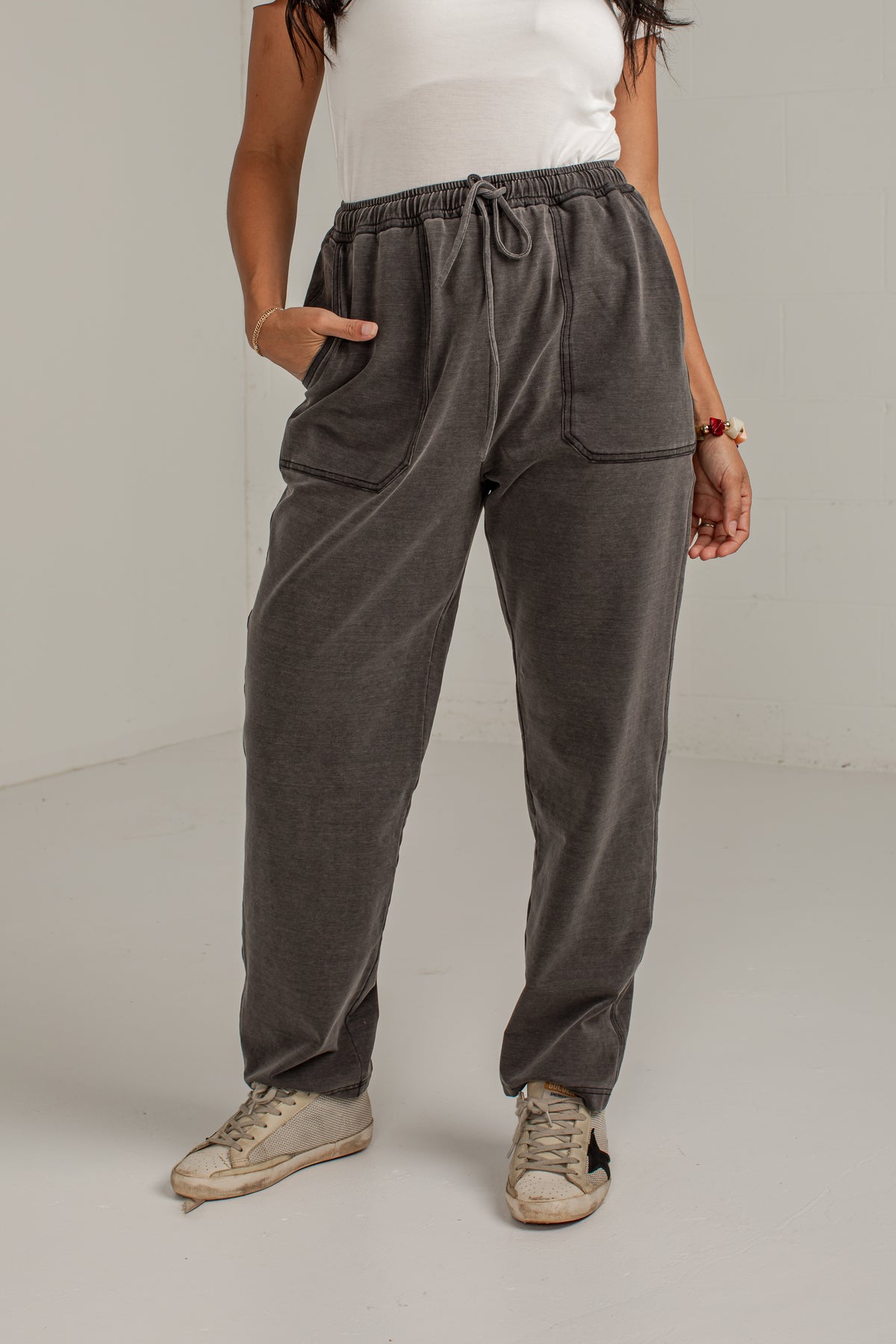 A person wearing dark gray drawstring sweatpants with side pockets, paired with a white sleeveless top and beige sneakers, standing against a plain light gray studio background.