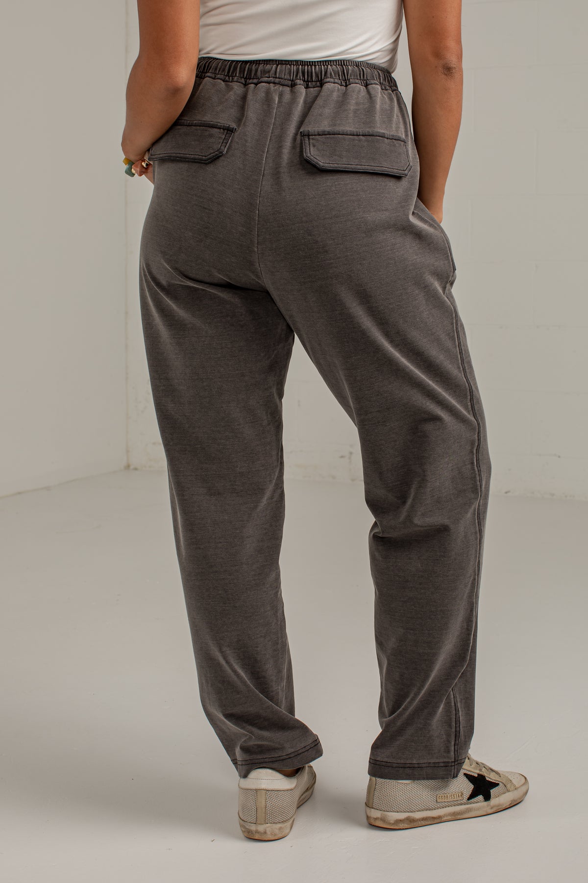 A person shown from the back wearing dark gray drawstring lounge pants with side pockets and a relaxed straight-leg fit, paired with a white top and light-colored sneakers.