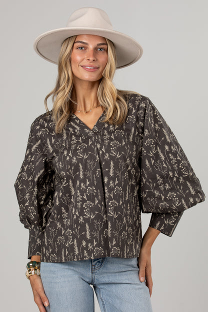 A woman wearing a dark brown and beige floral print blouse with voluminous sleeves, a wide-brimmed beige hat, and light blue jeans, photographed against a solid light gray background.