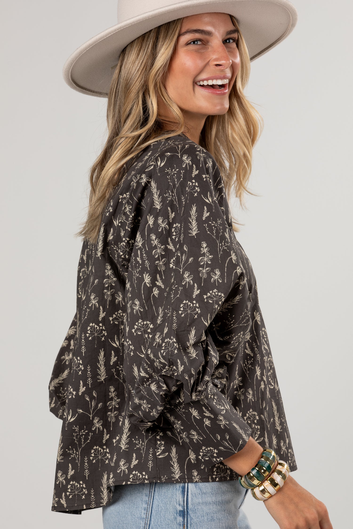 A woman wearing a dark brown long-sleeve blouse with a beige botanical floral pattern, styled with a wide-brimmed cream hat and light blue jeans against a light gray studio background.