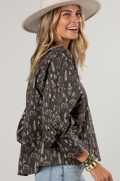 A woman wearing a dark brown long-sleeve blouse with a beige botanical floral pattern, styled with a wide-brimmed cream hat and light blue jeans against a light gray studio background.