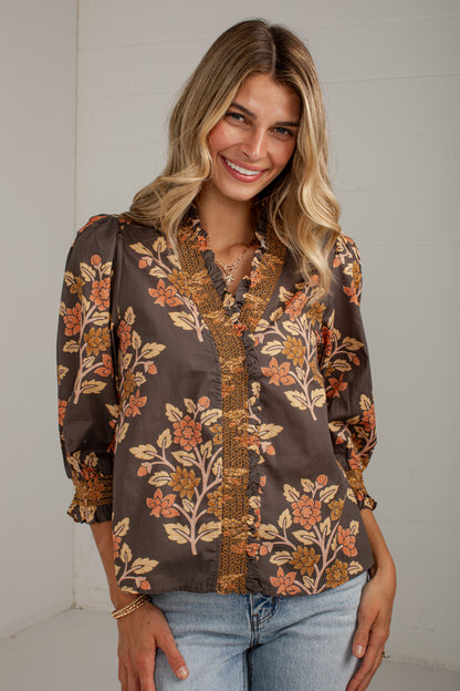 A woman wearing a brown long-sleeve button-up blouse with a floral pattern in orange, beige, and gold, paired with light blue jeans, standing against a white paneled wall.