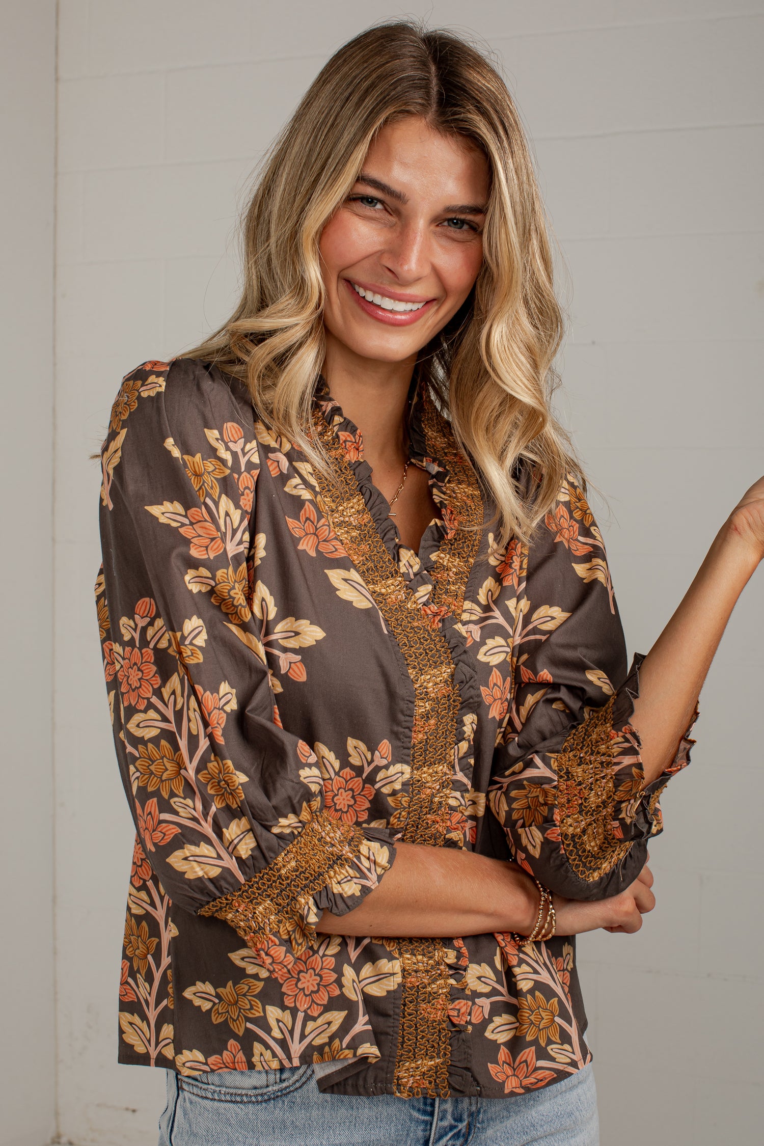 A woman wearing a brown floral blouse with orange and beige botanical patterns, paired with light blue jeans, standing against a white paneled wall.