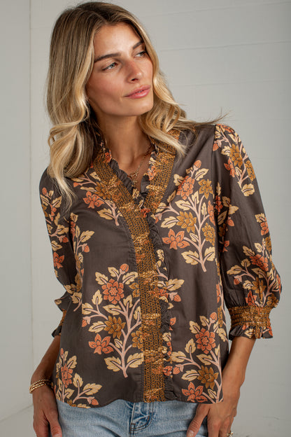 A woman wearing a brown long-sleeve blouse with a floral print in orange, beige, and brown, paired with light blue jeans, standing against a white paneled wall.