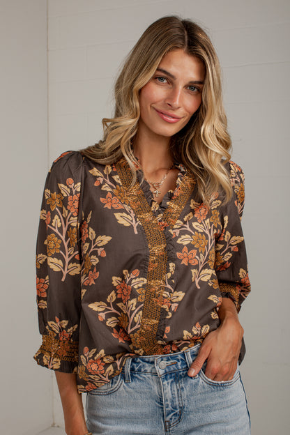 A woman wearing a brown floral blouse with gold and orange accents, paired with light-wash blue jeans, standing against a light gray wall.