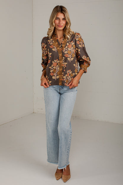 A woman modeling a brown long-sleeve blouse with a floral print in orange, beige, and green, paired with light-wash blue jeans and brown pointed-toe heels, standing against a white studio wall.