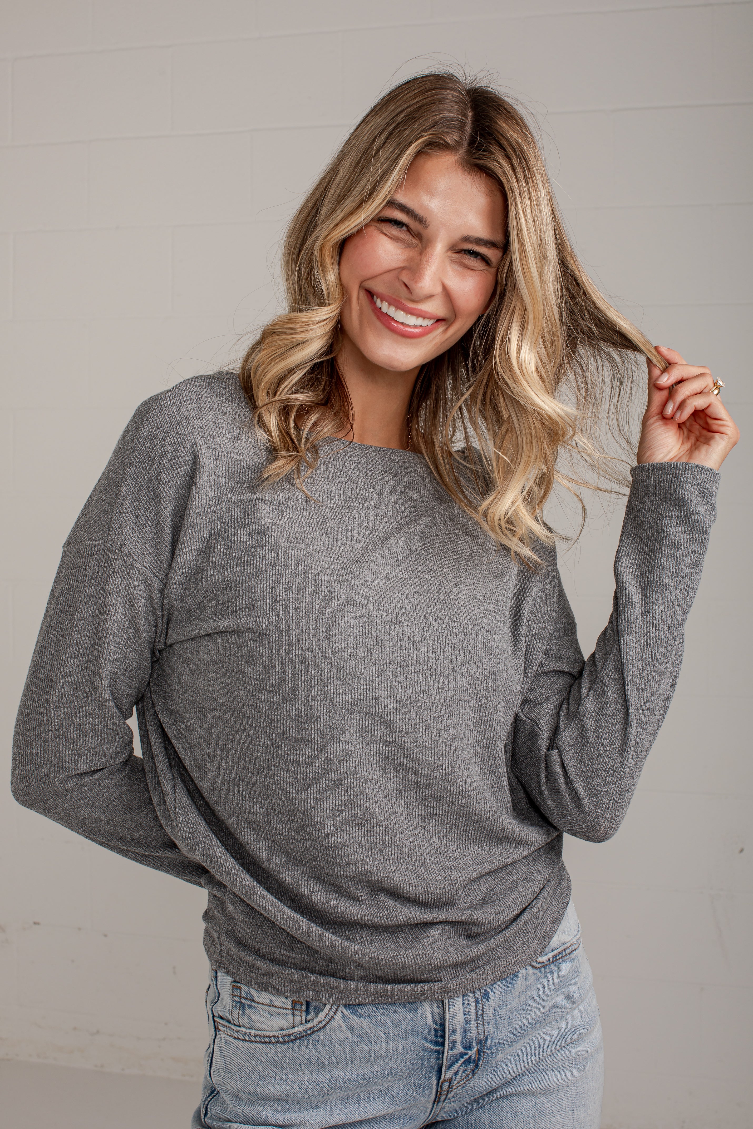 A smiling blonde woman wearing a heather gray long-sleeve crewneck sweater and light blue jeans, posing against a light gray paneled wall.