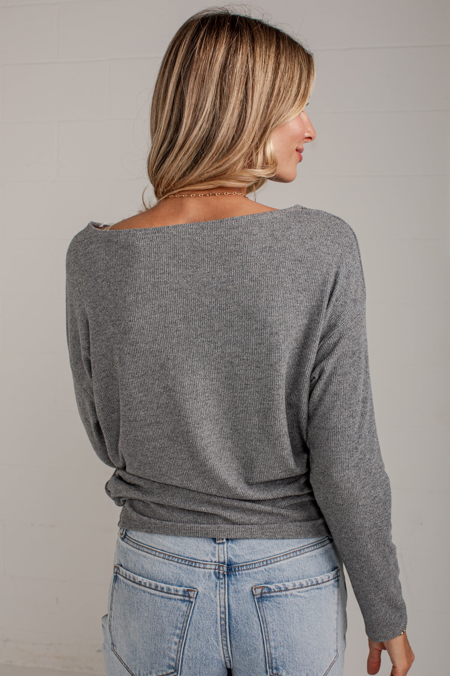 A woman shown from the back wearing a heather gray long-sleeve knit top with a wide boat neckline, paired with light blue jeans, against a light gray wall.